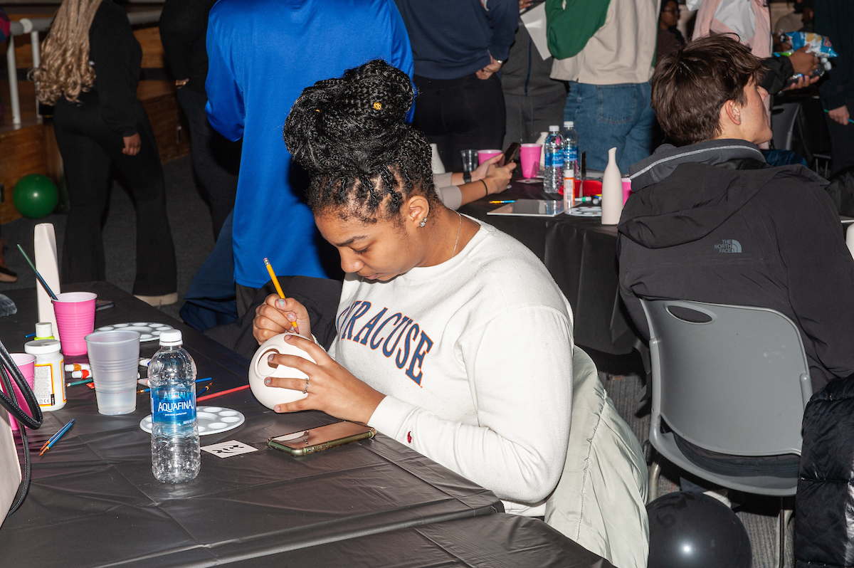 student in a Syracuse sweatshirt paints a round ceramic object seated at a table