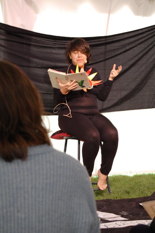 person sitting on a stool reading from a book to an audience
