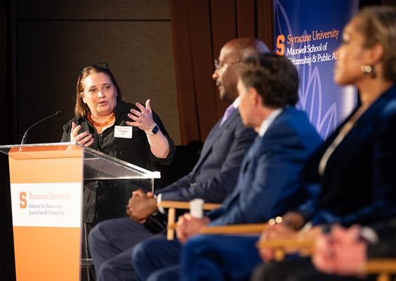 Margaret Talev speaks at a podium to panelists Van Jones, Bob Costas and Danielle Nottingham at an event in Los Angeles