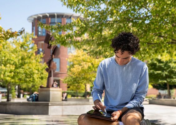 a student seated outdoors does work on a tablet