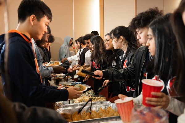 Various foods on a long table with students serving their peers on the other side of the table.