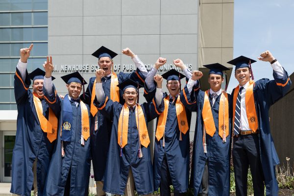 A group of graduates cheer in their Commencement regalia outside of the Newhouse School at Commencement 2023