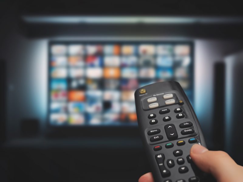 Hand holding a TV remote pointed at a blurred streaming service menu on a screen