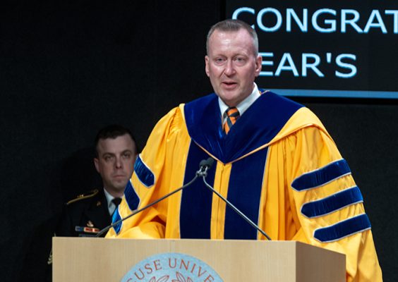 Individual standing at podium speaking while wearing a gold colored graduation robe.