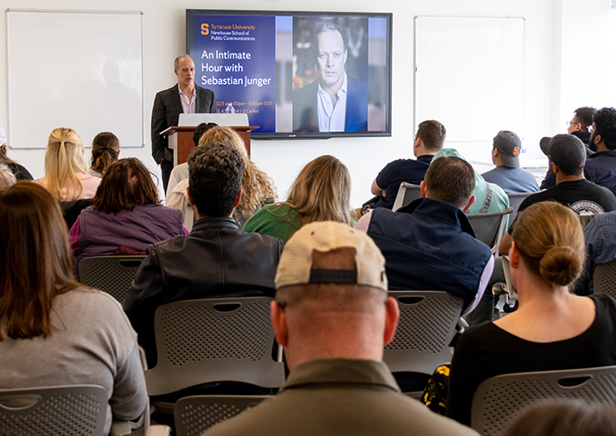 Man standing at a podium speaking to an audience with a powerpoint presentation on the wall behind him.