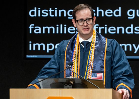 Man standing t podium speaking wearing navy blue graduation gown and gold tassels.