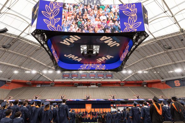 JMA Dome Dome during Commencement. Students in caps and gowns are standing on the floor with the stage and a view of the giant screen above the field with the audience on the screen.
