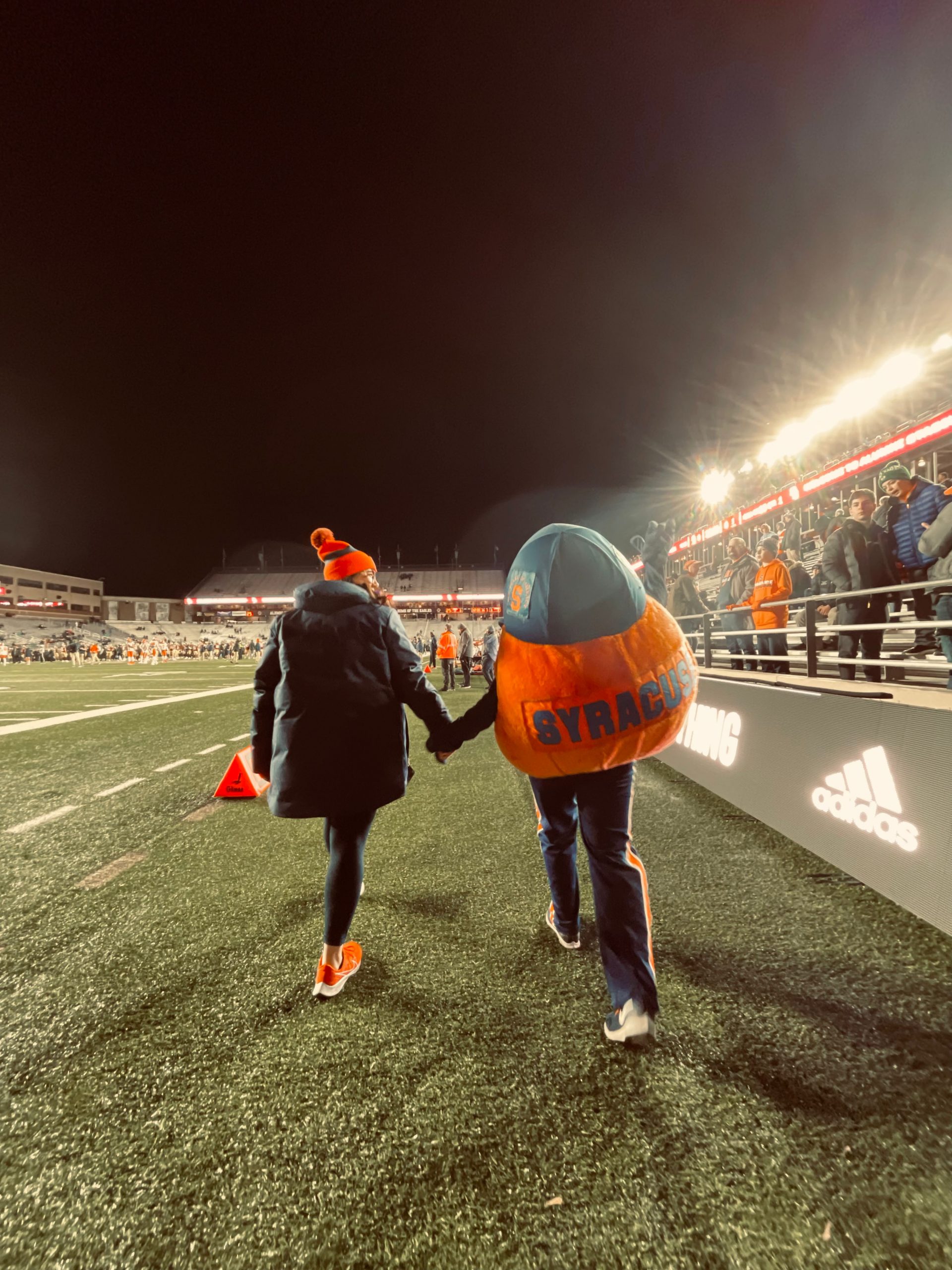 Otto and an individual holding hands walking away from the camera down a football field