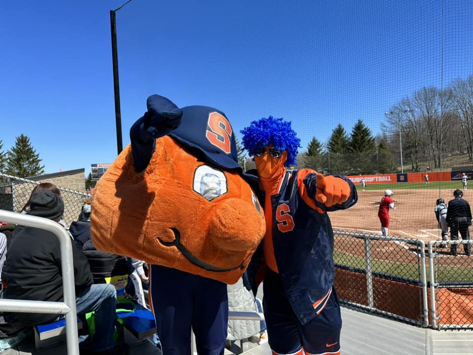 Individual dressed head to toe in Syracuse Athletics gear standing with Otto at a softball game.
