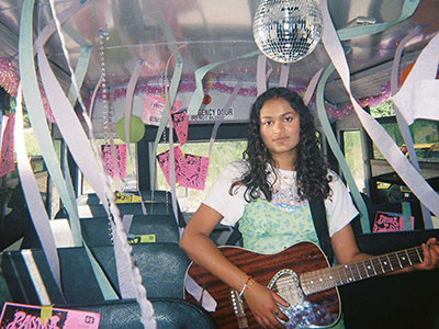 Individual standing inside of a bus decorated with streamers and a disco ball holding a guitar.