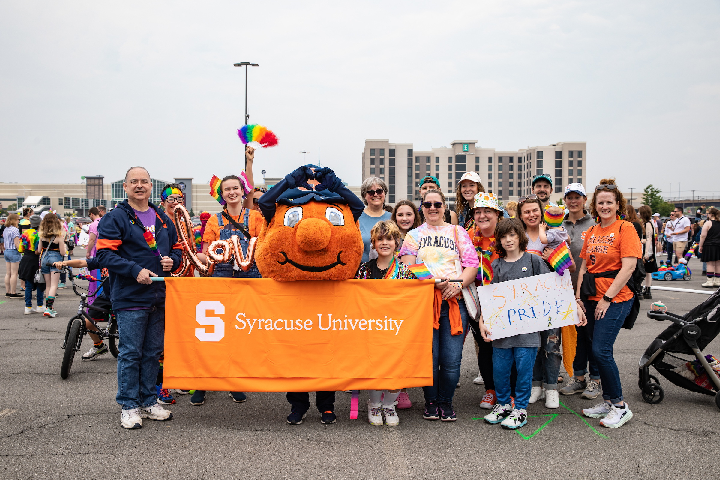 Group of people standing together with Otto the Orange and an orange Syracuse University banner before the Pride parade. 