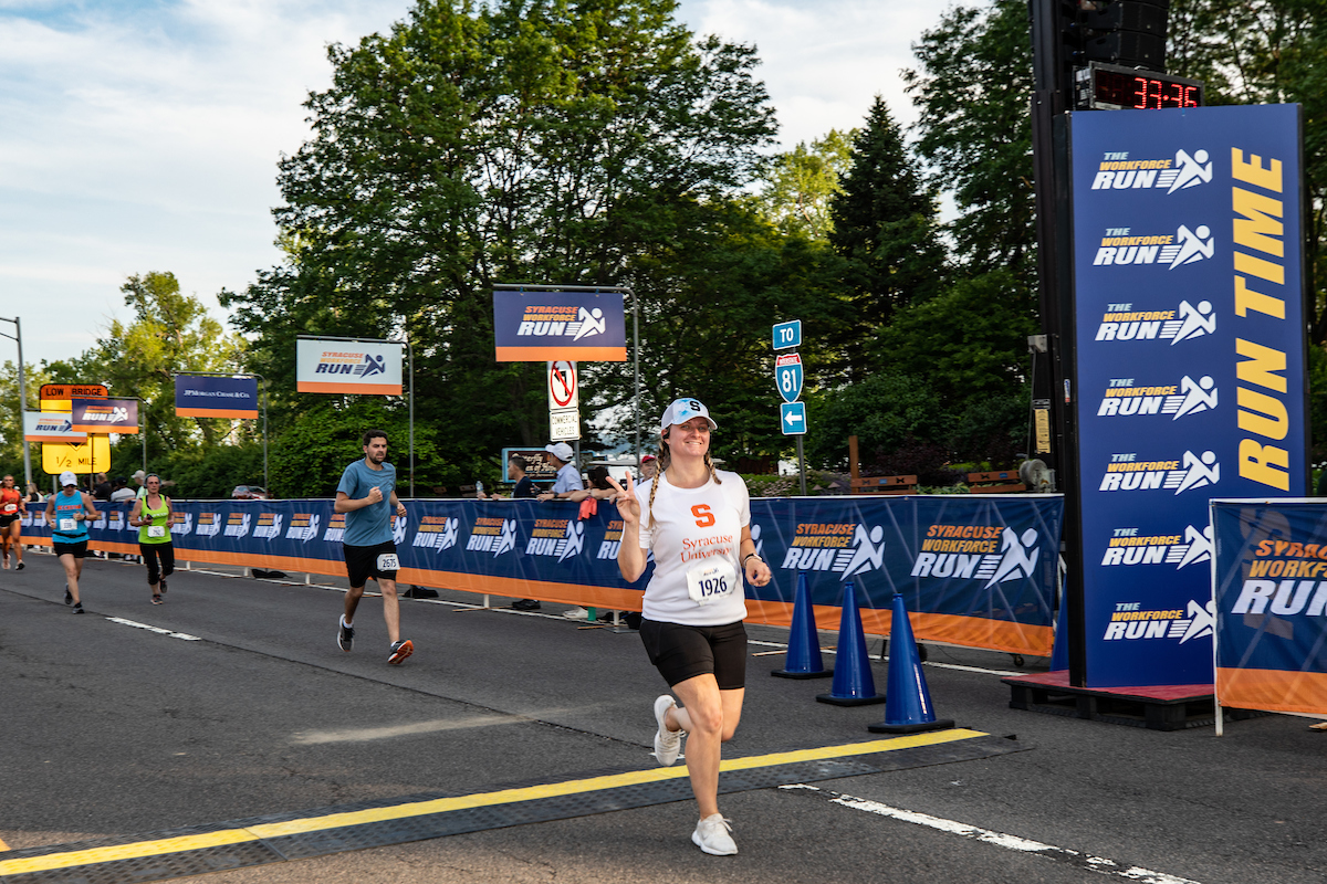Individual in a Syracuse University t-shirt flashes the peace sign as they cross the finish line at the Syracuse WorkForce Run