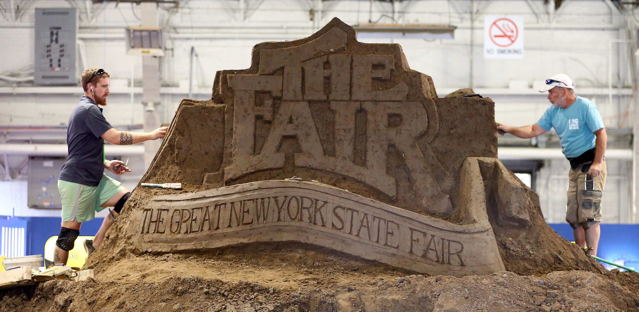 Sand sculpture of the Great New York State Fair logo with two individuals working on it. 