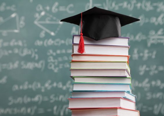 books stacked up in front of a blackboard with a mortarboard hat on top