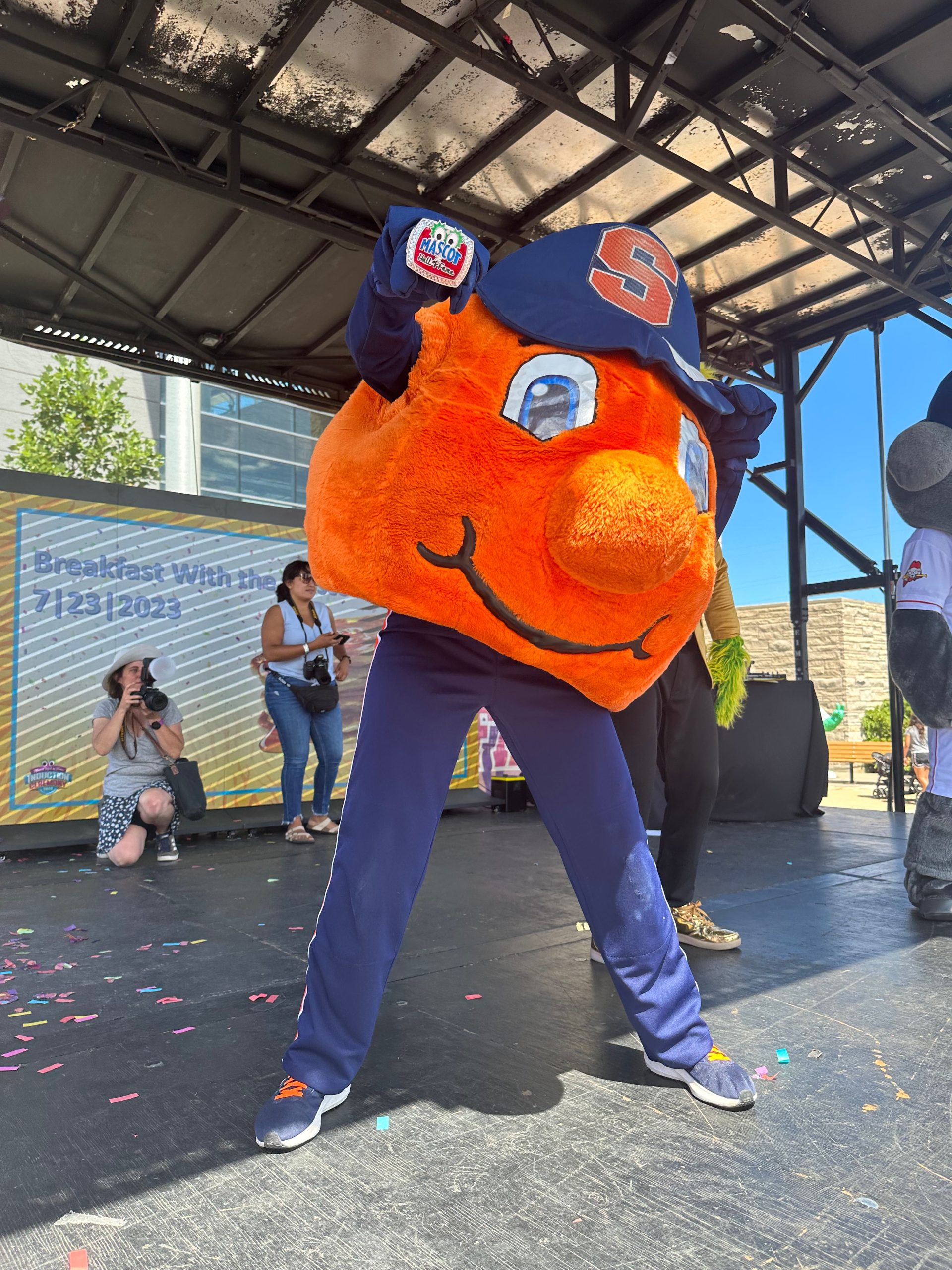 Otto the Orange standing on a stage showing off their hall of fame ring 