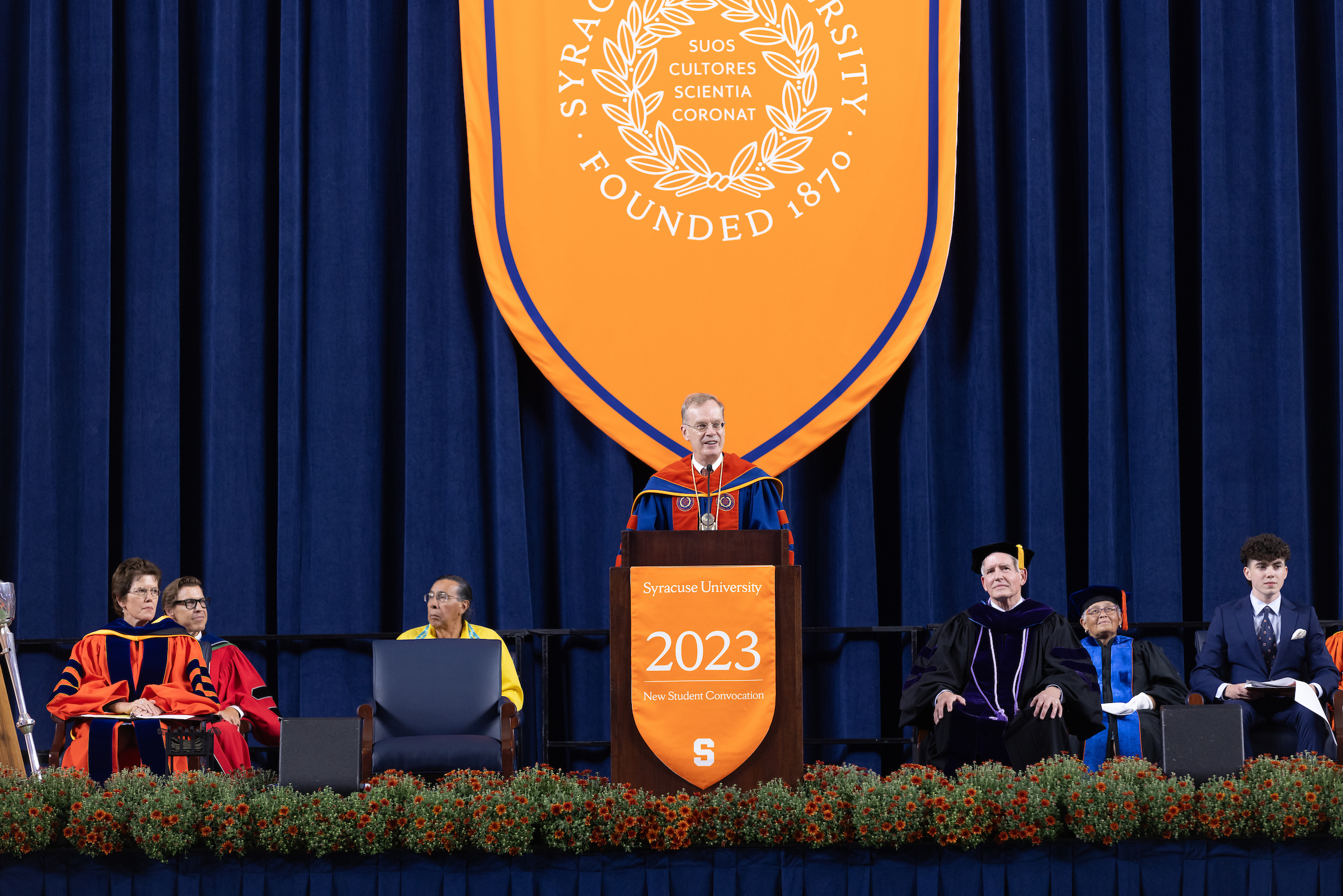 Chancellor standing at the podium speaking on stage with other sitting on the stage nearby