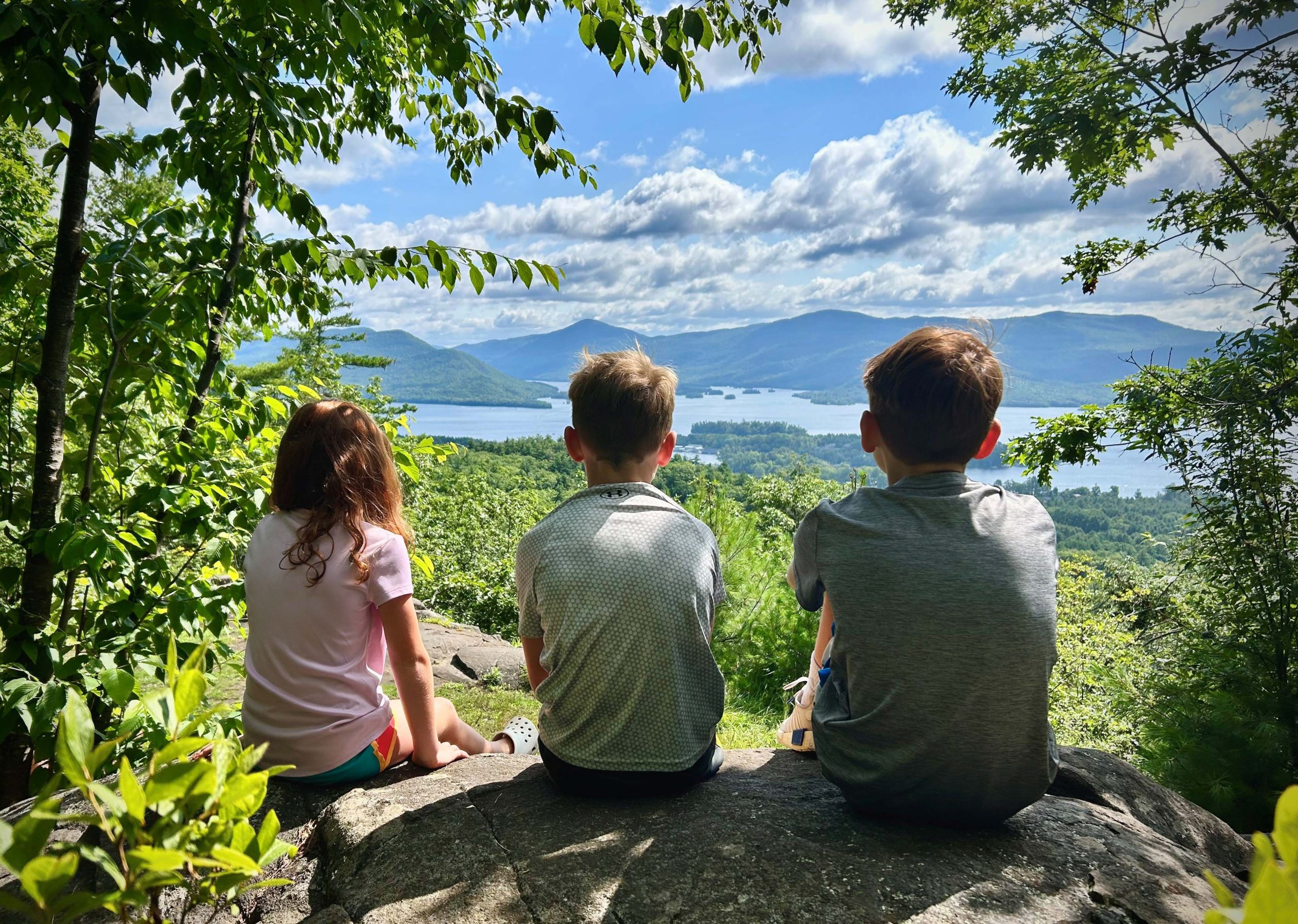 Three children sitting on a rock with their backs to the camera overlooking a lake