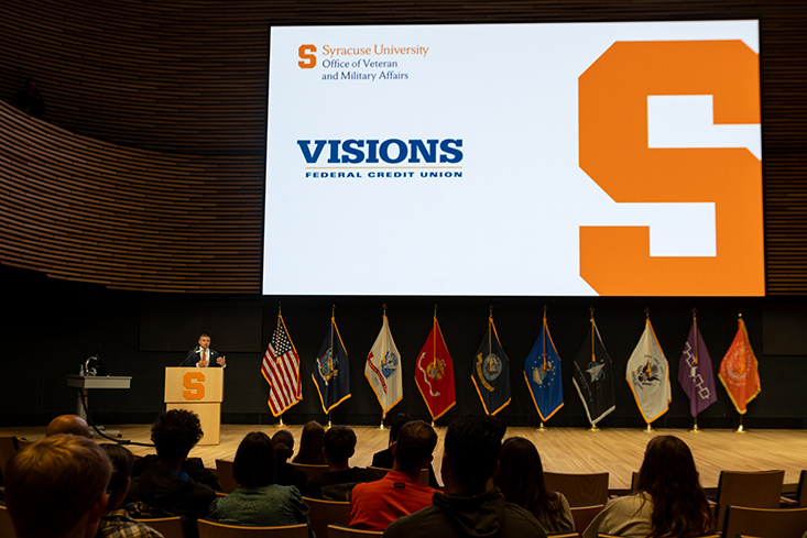 Individual standing at a podiium on a stage speaking with the presentation on the wall behind them with a Syracuse University and Visions Federal Credit Union logo on it.