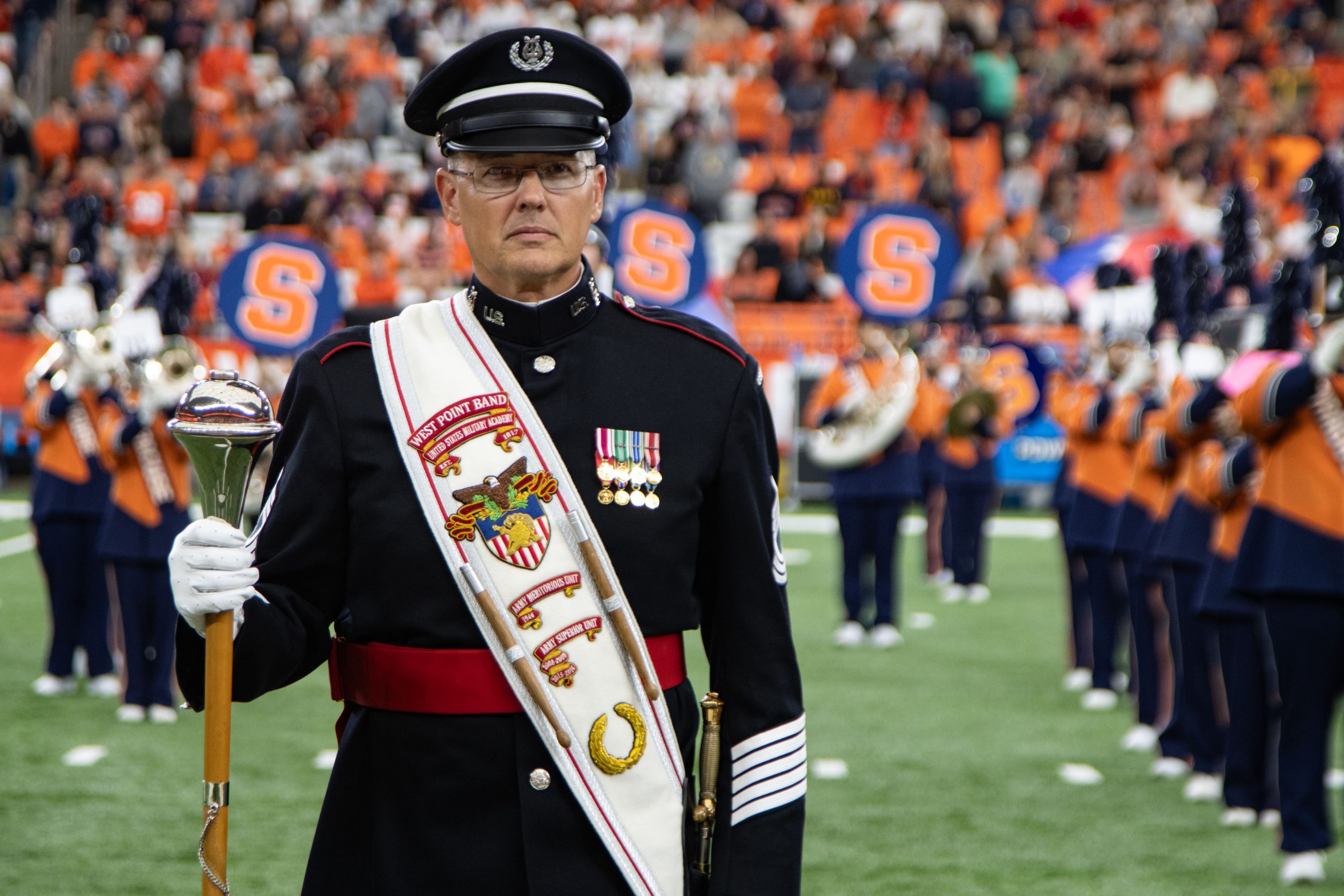 Individual standing in full military uniform on the field in the Dome with the University's marching band in the background. 