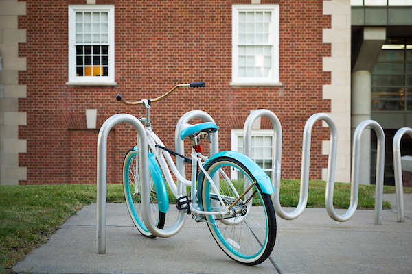 A turquoise bicycle is locked up at a bike rack with the West Facade of Maxwell filling the background.