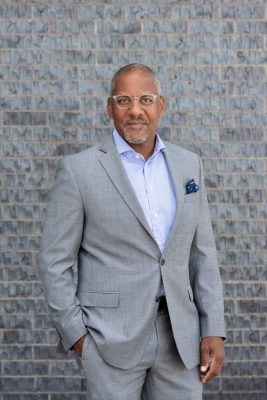 Person in a light gray suit and striped shirt standing against a gray brick wall, with one hand in pocket and a blue pocket square.