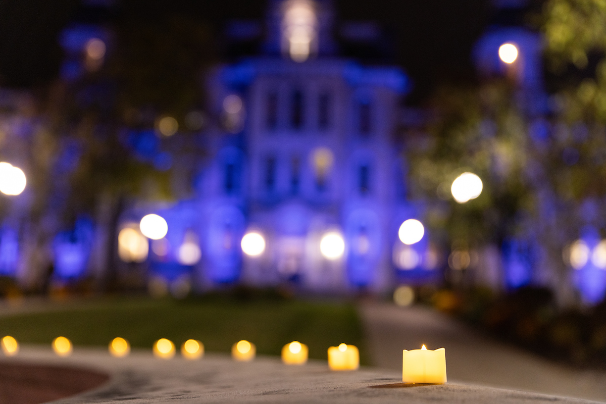 Candles on the Remembrance Wall at nighttime with the Hall of Languages lit up in blue in the background.