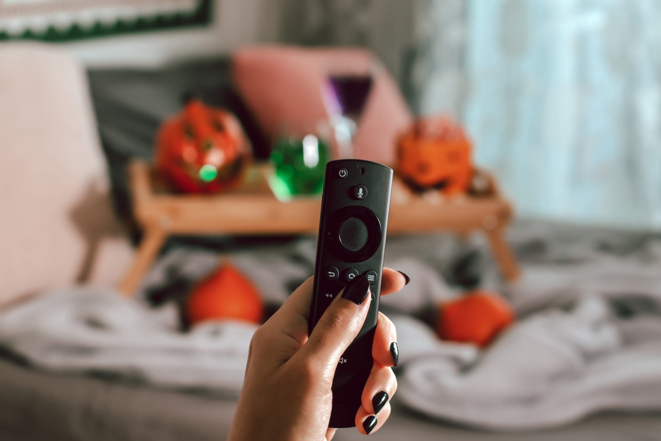 Hand holding a tv remote with a pillow and tray table in the background decorated with Halloween decorations.