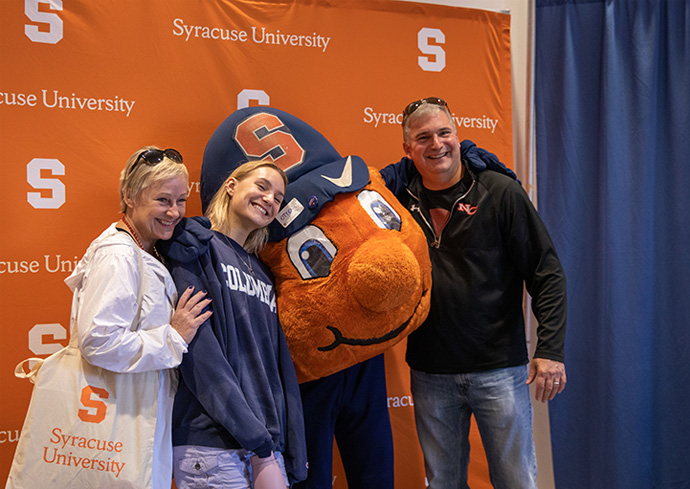 a student and their parents pose with Otto in front of a Syracuse University banner
