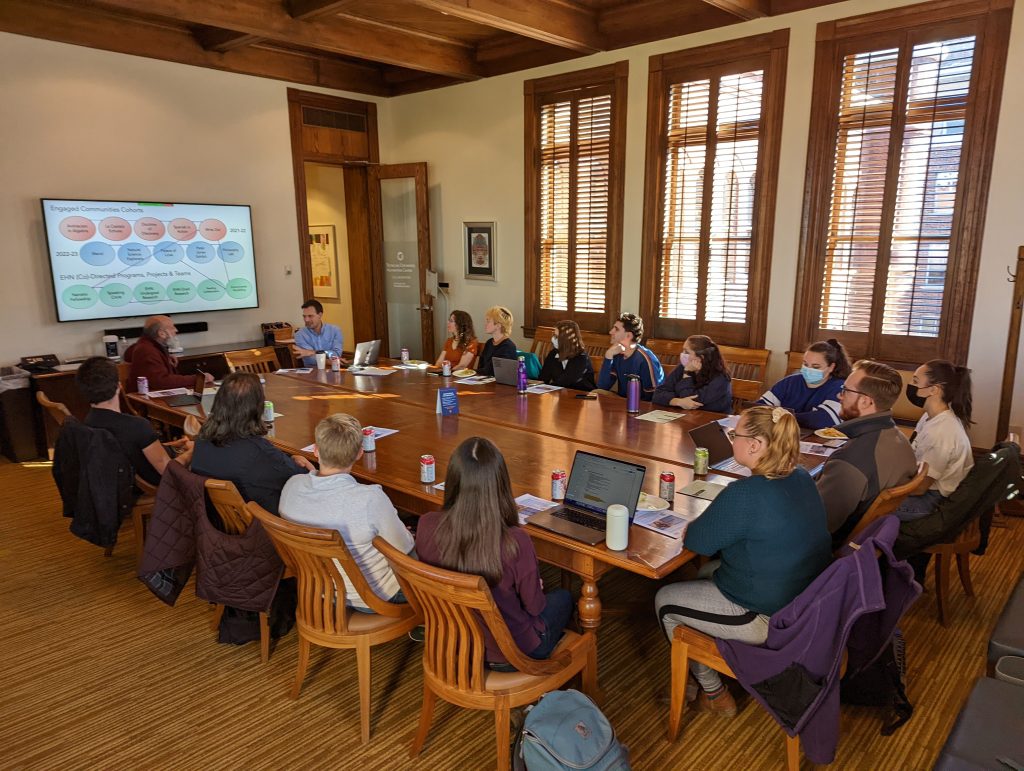 Large group of people sitting around a table with a screen in the front of the room with a presentation of it.