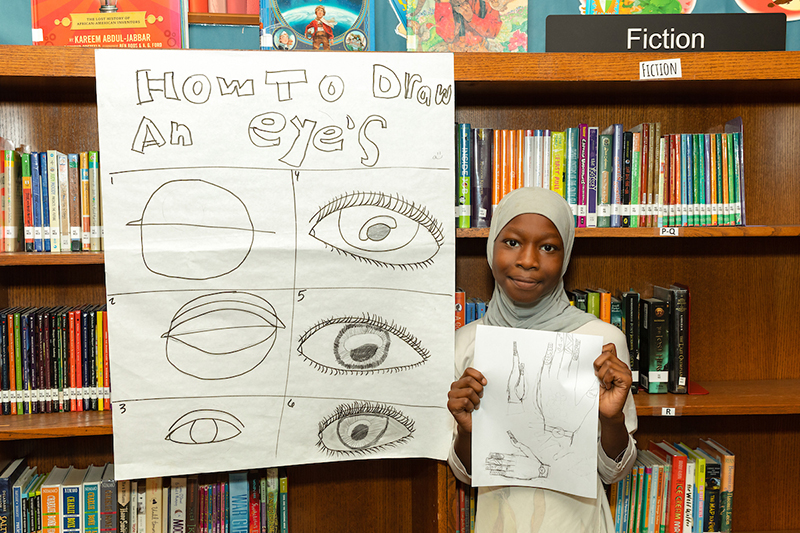 elementary school student standing in front of a bookcase shows off her project detailing how to draw hands and eyes