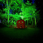 Sppoky pumpkin shaped house in the dark with trees behind it lit up in green.