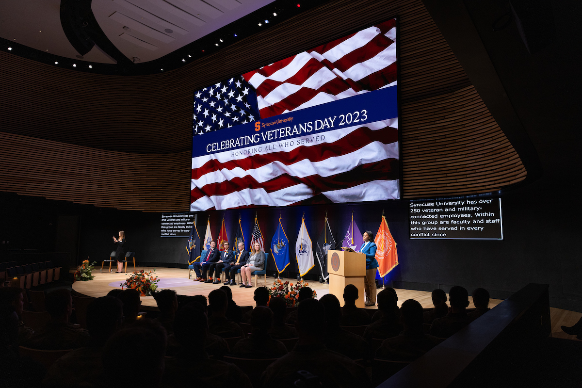 Auditorium with a stage and a large American flag on the screen. People sitting in chairs on the stage and various flags lining the stage under the screen. 