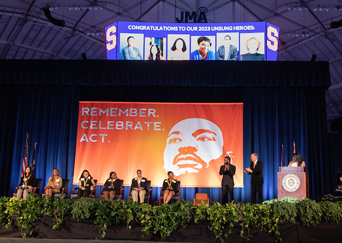 people sitting and standing on stage in front of large curtain that states Remember. Celebrate. Act. with image of Martin Luther King Jr.