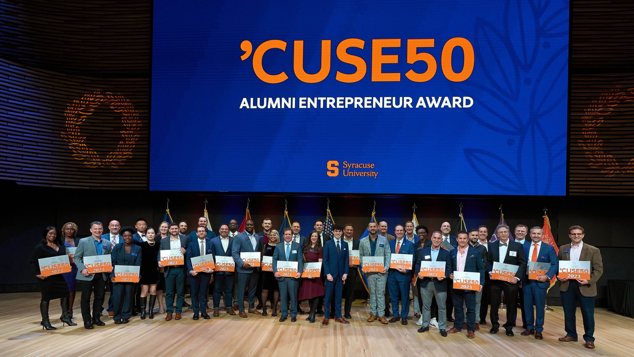 Large group of people on a stage with a large blue screen that says ’CUSE50 Alumni Entrepreneur Award