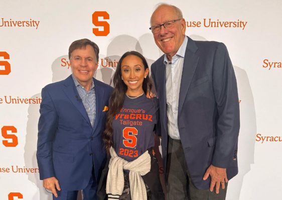 Three Syracuse University alumni pose for a photo in front of a white backdrop with an Orange block S and the words Syracuse University.