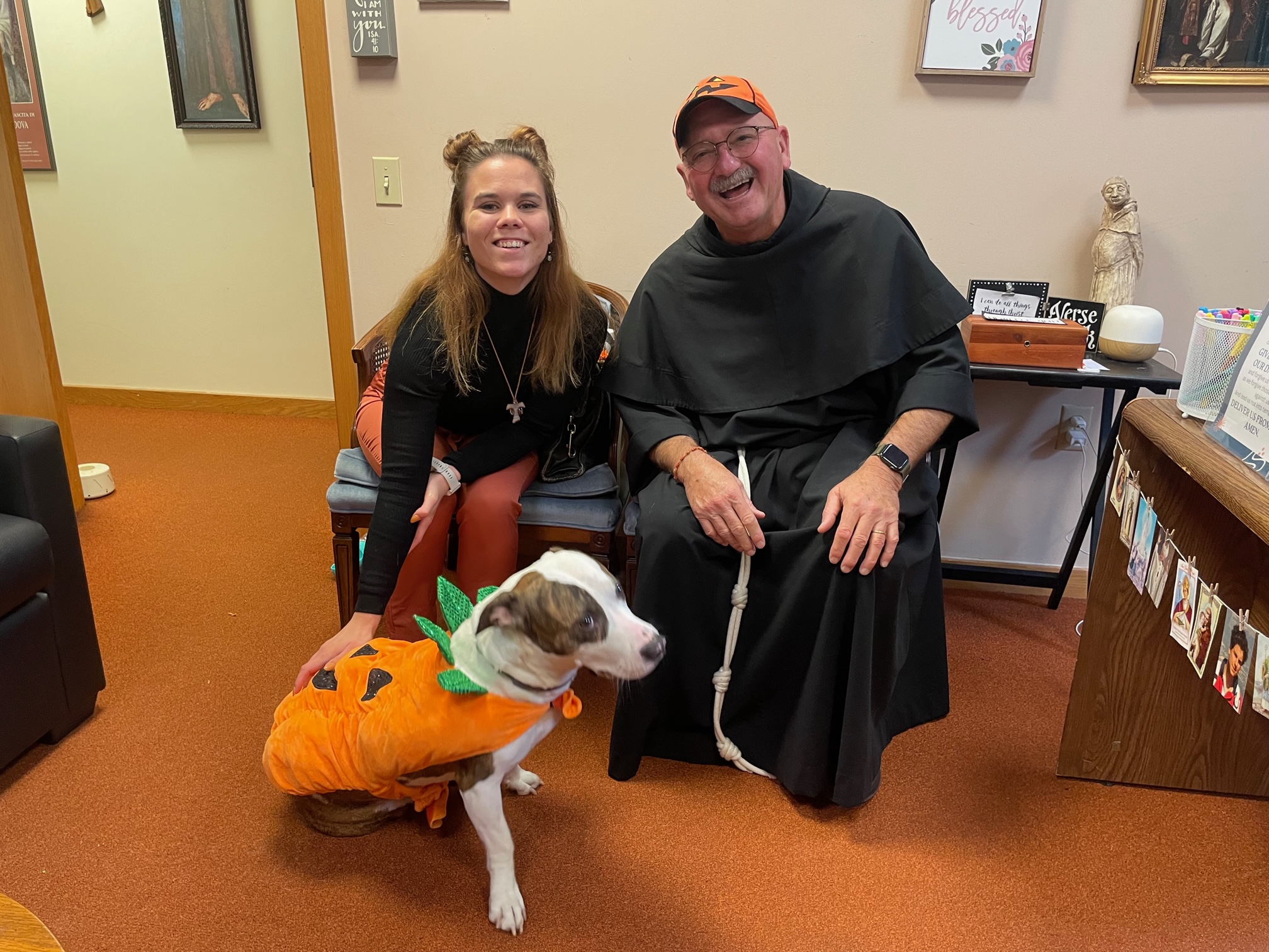 Two people sitting in chairs with a dog in a Halloween costume.