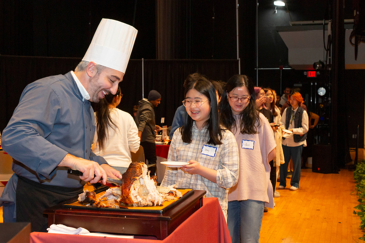 Man with a chef's hat carves a turkey and serves it to a line of students.