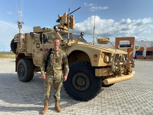 A man poses for a photo in military gear in front of an armored vehicle.