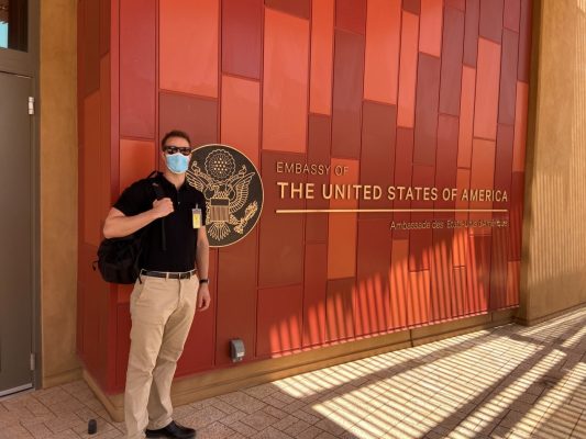 A man wears a mask while posing for a photo in front of the U.S. Embassy.