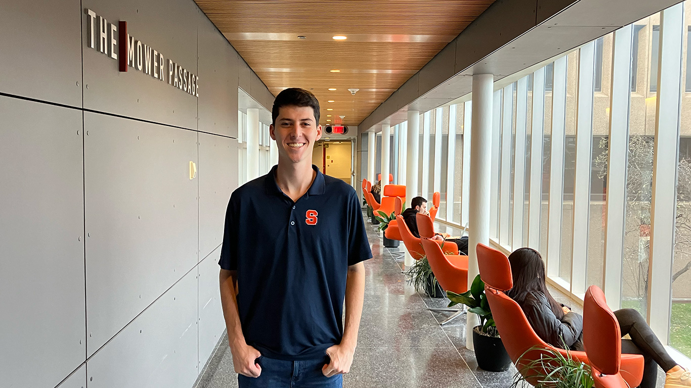 student Adam Avin stands in a hallway smiling at Newhouse