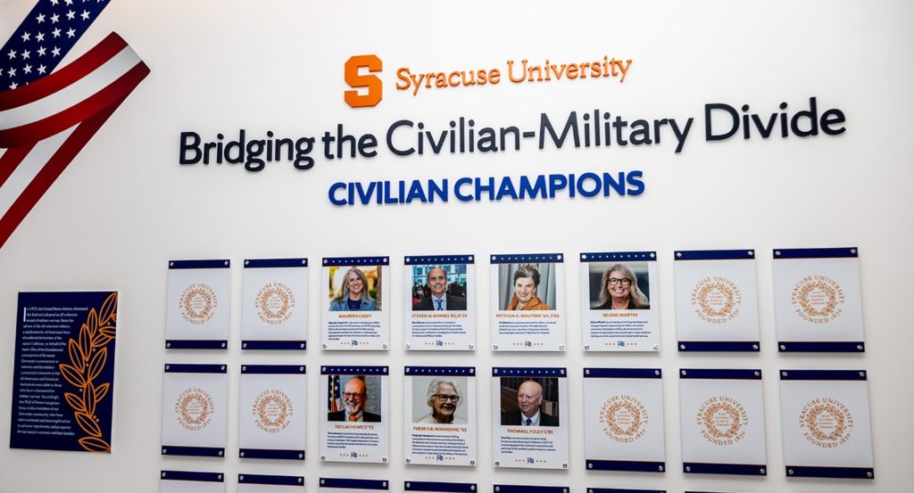 Wall display at Syracuse University titled “Bridging the Civilian-Military Divide,” featuring framed photos of Civilian Champions and a partial American flag.