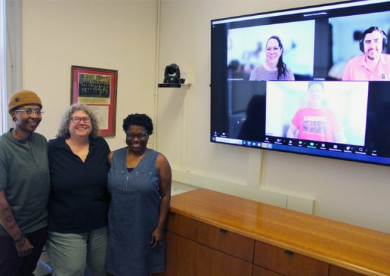 three individuals stand together with three people displayed on a Zoom screen