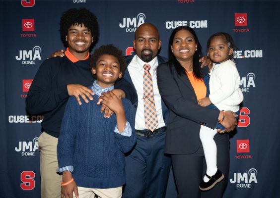 A husband and wife pose for a photo with their two sons and daughter in front of a blue step and repeat banner