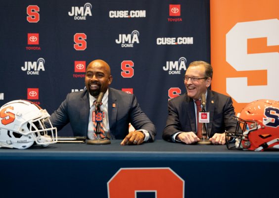 Two men answer questions at a podium. A white Syracuse football helmet is on the left of the podium and an Orange Syracuse football helmet is on the right.