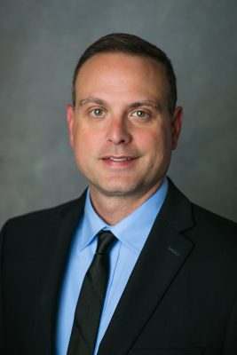 Person in a black suit and light blue shirt against a neutral gray background.