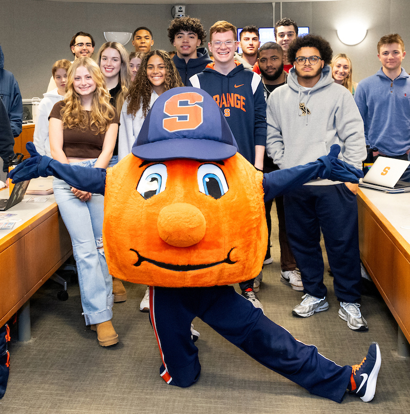 Otto the Orange posing with a group of students in a classroom.