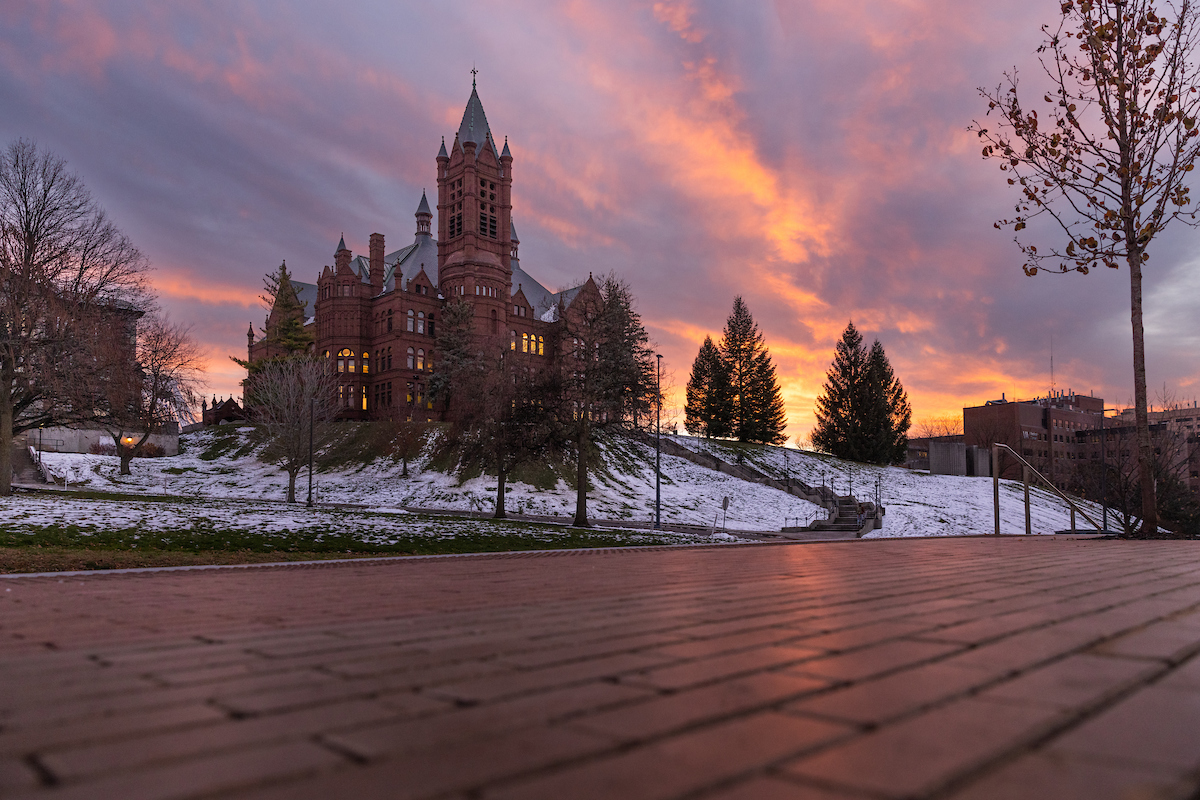 View of the promenade with snow on the ground and Crouse College in the background with an orange and purple sunset. 