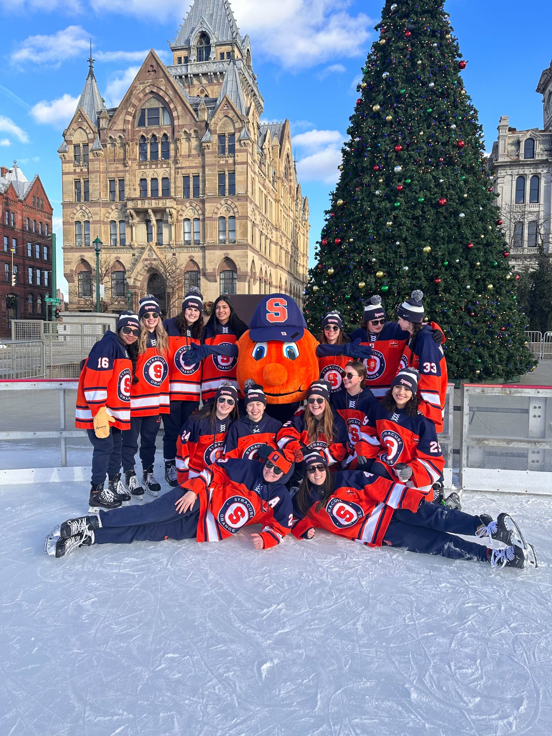 Womens ice hockey team posing with Otto the Orange on the rink at Clinton Square with the Christmas Tree in the background