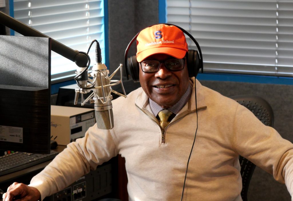 A man wearing an Orange Syracuse University hat poses for a photo in front of a microphone.