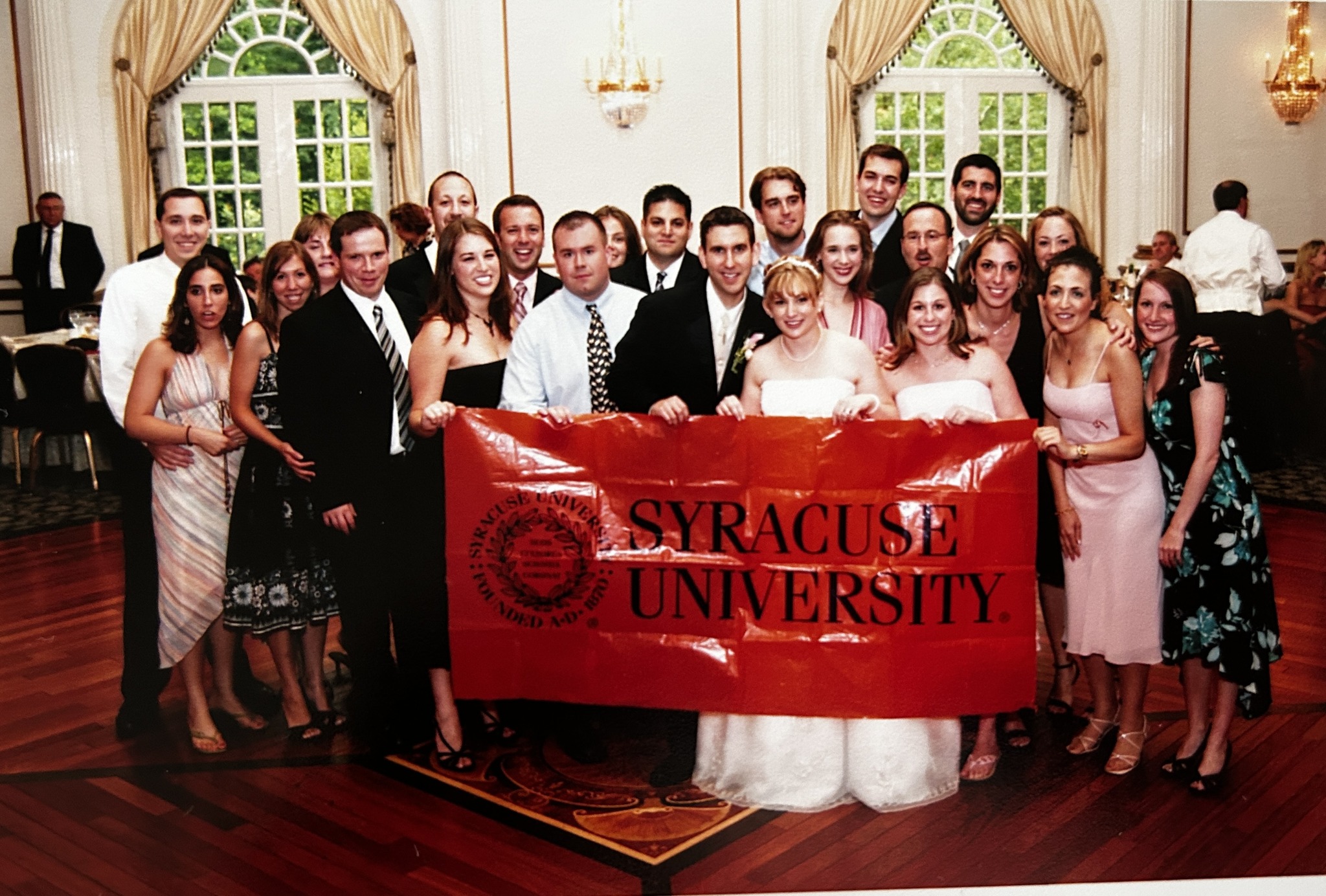 Large group of people standing together at a wedding holding a Syracuse University banner